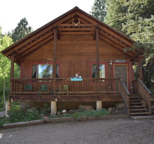 A wooden cabin with a covered porch, set among trees, with a gravel driveway in front. The porch has green chairs, potted plants, and a sign above the door. At Vallecito Lake, Colorado.